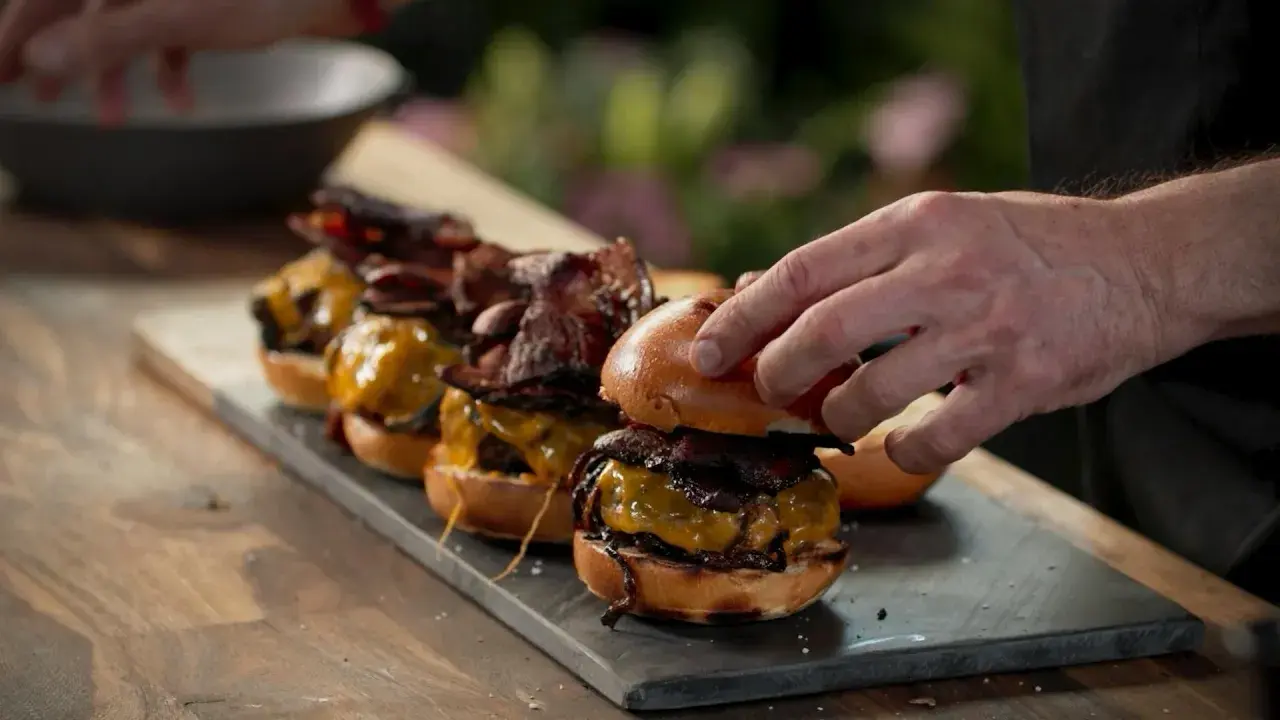 En hand lägger på toppen av en smashed burger med ost och bacon. Flera likadana burgare står uppradade.