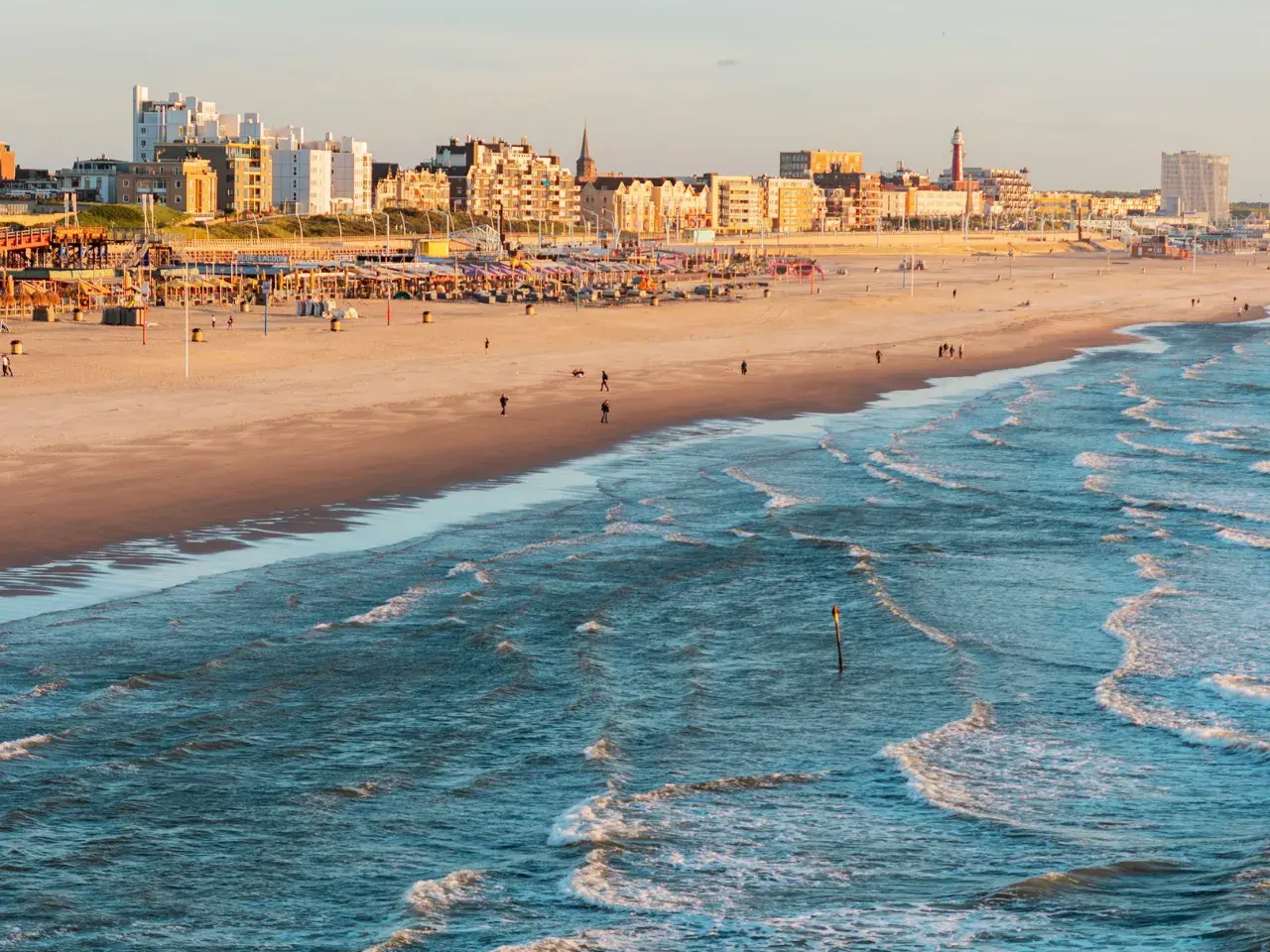 Ein sonniger Tag an der Nordsee Holland. Wellen brechen am Strand, im Hintergrund die Stadtkulisse mit Hotels und Strandpavillons.