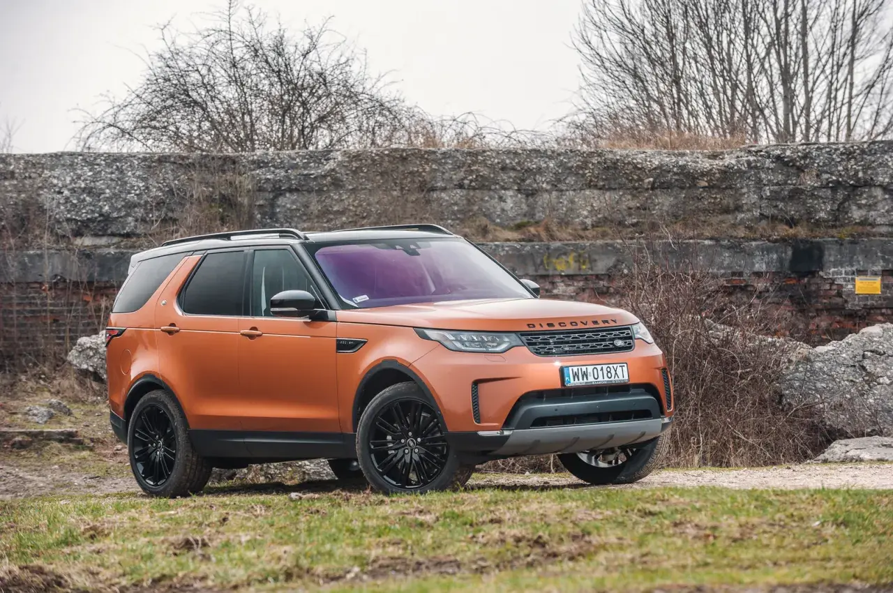 Pomarańczowy Land Rover Discovery 5 z czarnymi felgami stoi na trawiastym podłożu, na tle kamiennego muru i suchych krzewów.