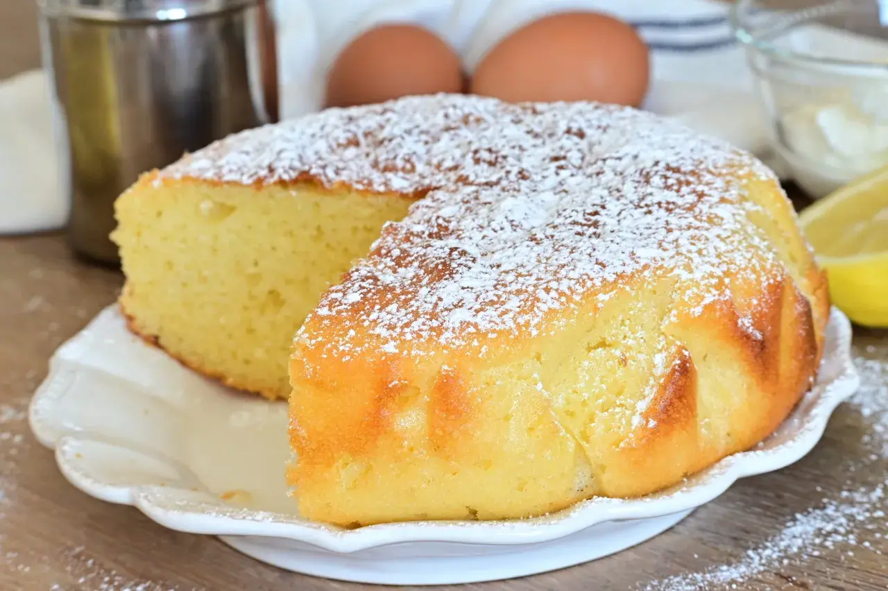 Una soffice torta al limone, spolverata di zucchero a velo, pronta per essere gustata. Ingredienti freschi come uova e limone sullo sfondo.