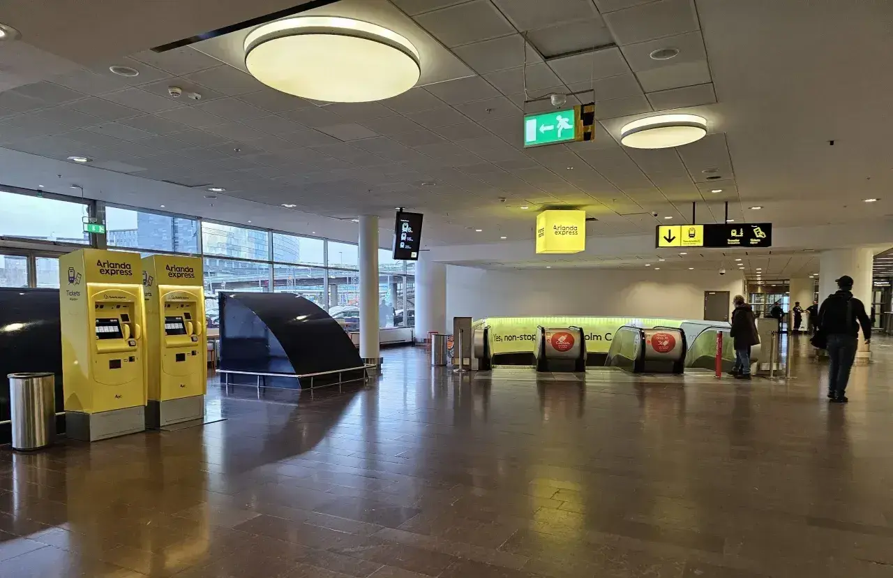 Żółte automaty z biletami na Arlanda Express na sztokholmskim lotnisku. W tle schody ruchome i znaki kierunkowe.