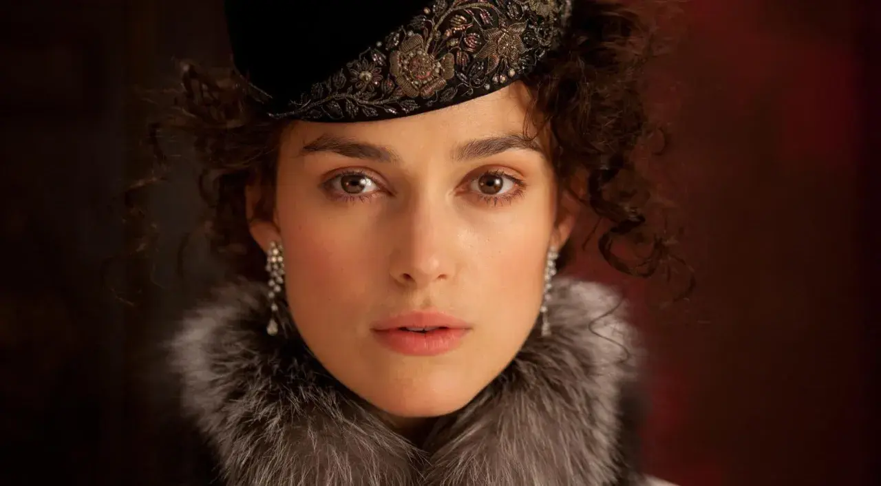 Anna Karenina w eleganckim futrze i ozdobnym kapeluszu, z błyszczącymi kolczykami, patrzy wprost przed siebie.