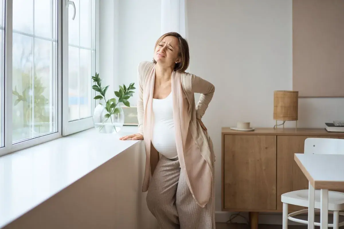 Gravid kvinna med ont i svanskotan lutar sig mot ett fönster. Hon håller handen på ryggen och grimaserar av smärta.