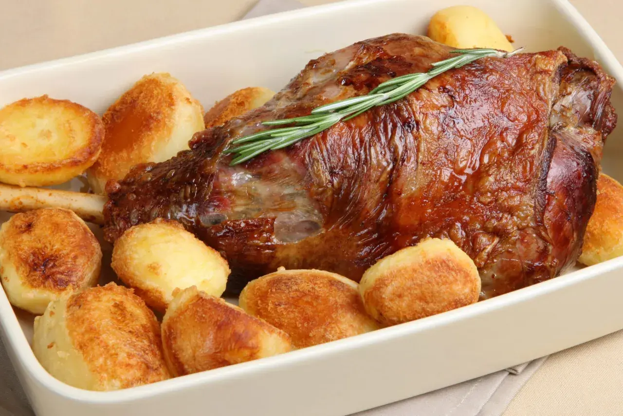 Gigot d'agneau rôti, garni de romarin, entouré de pommes de terre au four dorées et croustillantes. Un plat savoureux prêt à être dégusté.