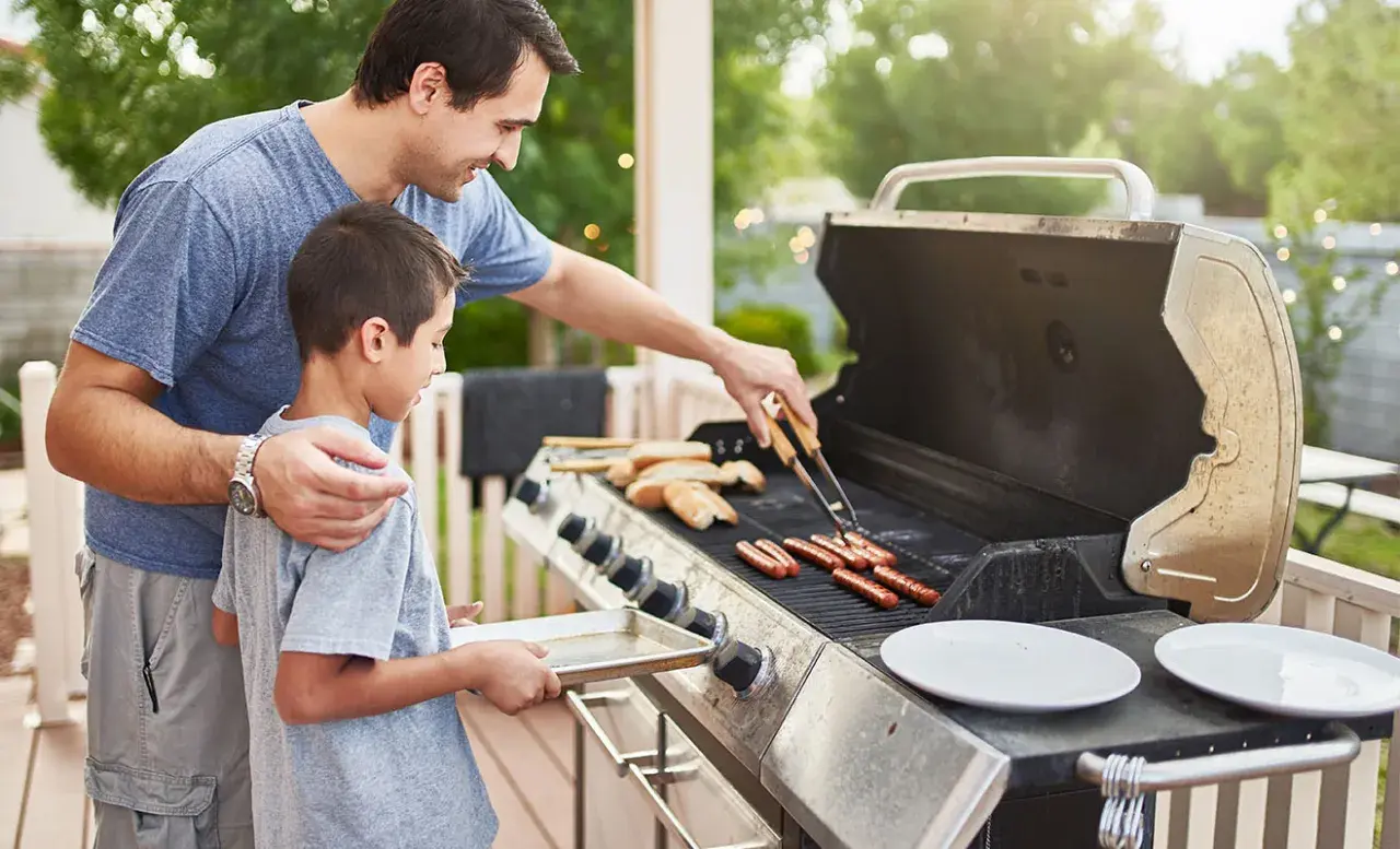 Un père et son fils préparent des saucisses sur un barbecue à gaz. Le père utilise des pinces pour retourner les saucisses pendant que le fils tient un plateau.