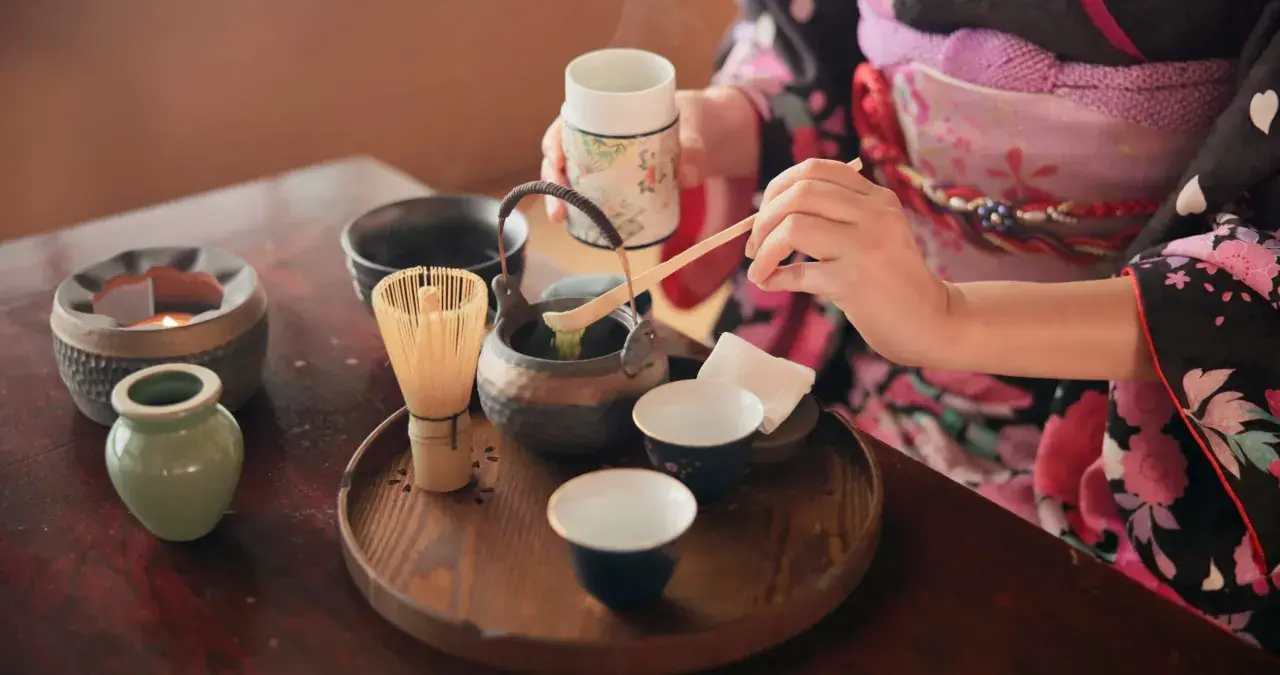 Eine Person in einem Kimono bereitet eine traditionelle matcha zeremonie vor. Ein Chasen (Bambusbesen) und eine Schale mit Matcha-Pulver sind auf einem Holzteller arrangiert.