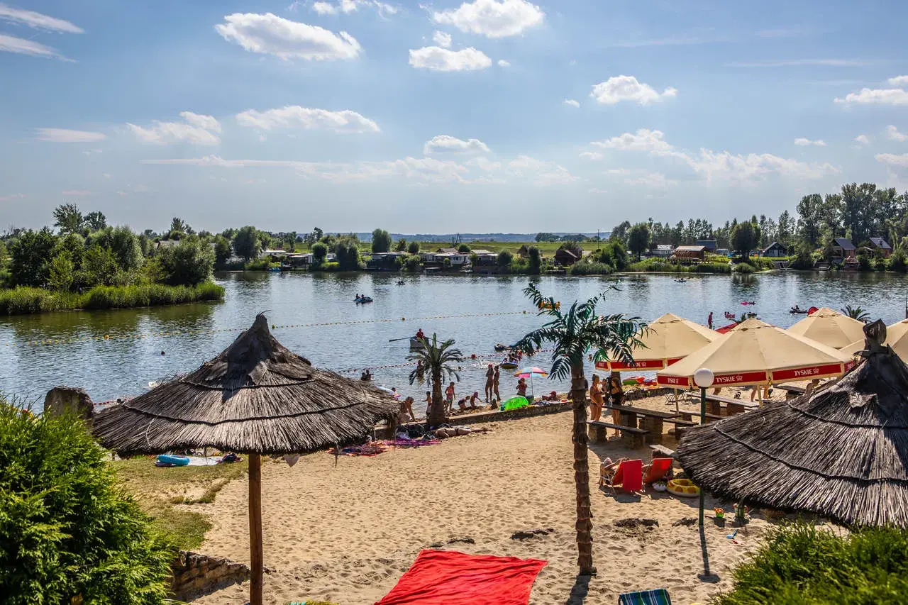 Słoneczny dzień na plaży w Kuźni Raciborskiej. Ludzie odpoczywają pod parasolami, kąpią się w jeziorze i pływają łódkami.