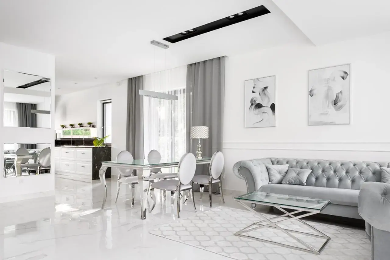 Eleganckie, nowoczesne wnętrze w stylu glamour. Błyszczące płytki, szara sofa i szklany stół tworzą luksusową atmosferę.