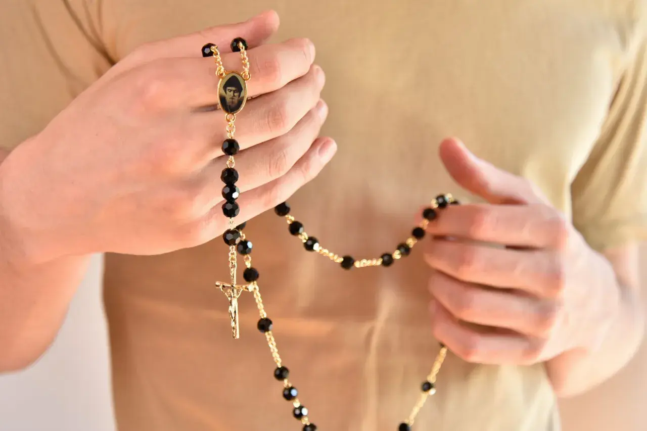 Dłonie trzymają złoty różaniec z czarnymi paciorkami i medalikiem. Zastanawiasz się, ile kosztuje różaniec w Watykanie?