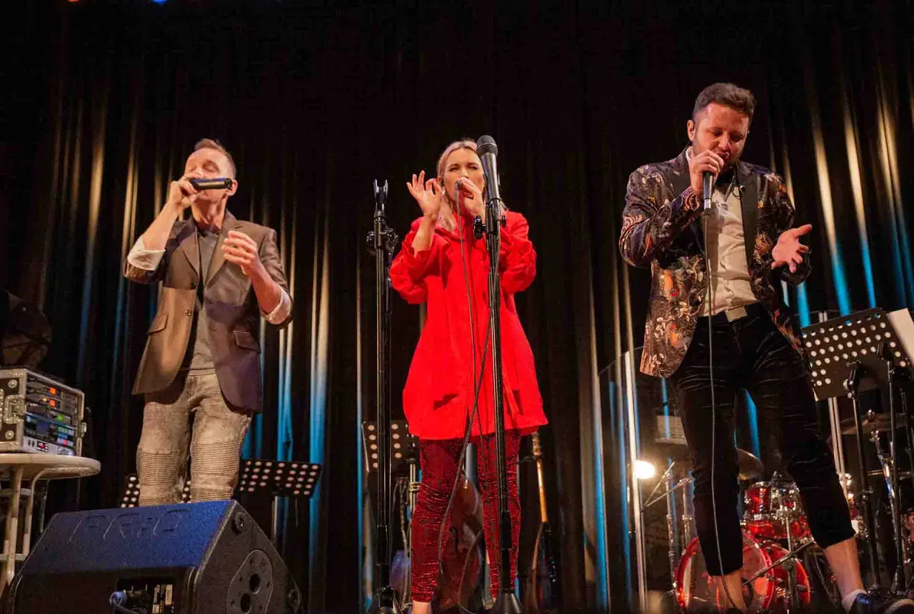Trzech wokalistów na scenie podczas warsztatów wokalnych. Kobieta w czerwieni, mężczyzna w marynarce z wzorami i mężczyzna grający na harmonijce.