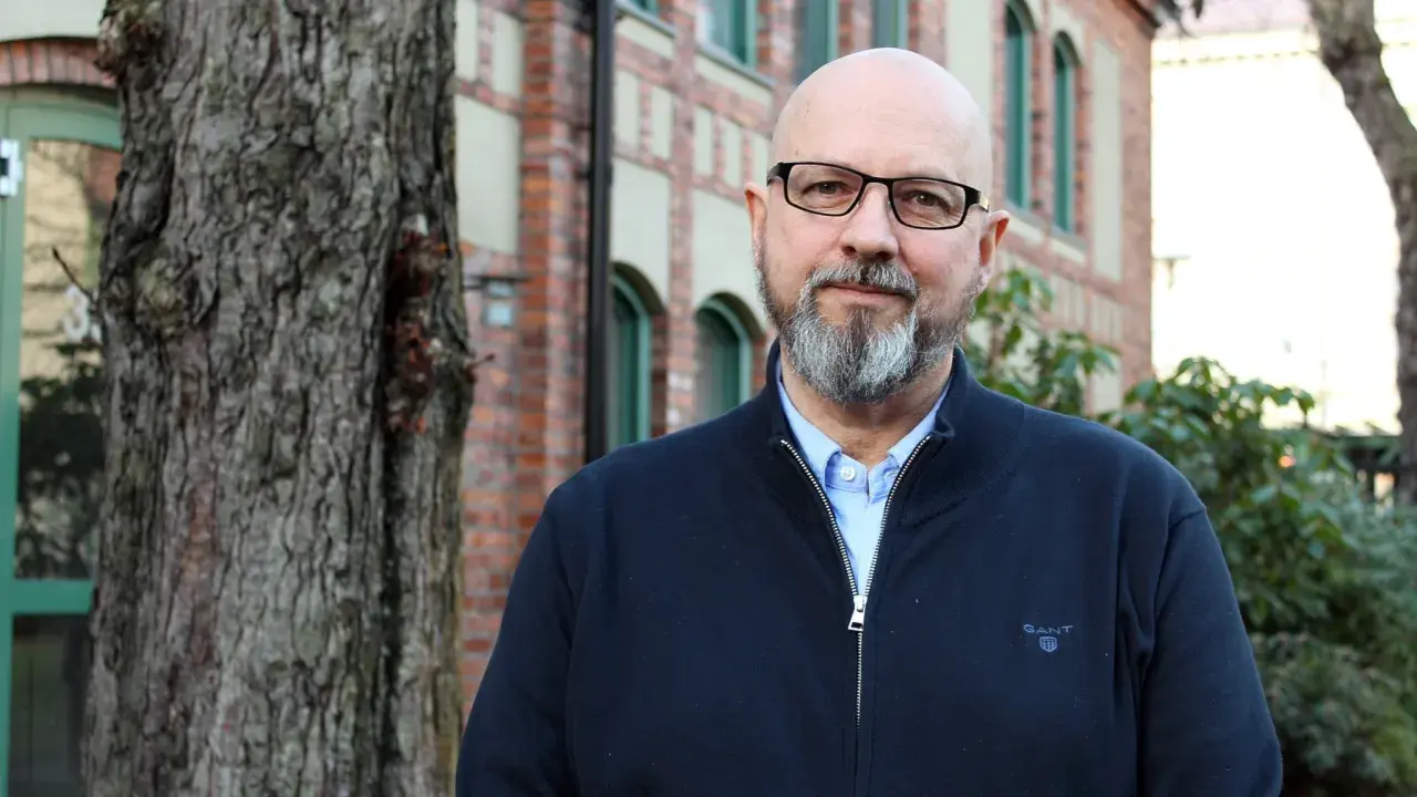 Kaj Stenman, en man med helskägg och glasögon, står framför en tegelbyggnad.