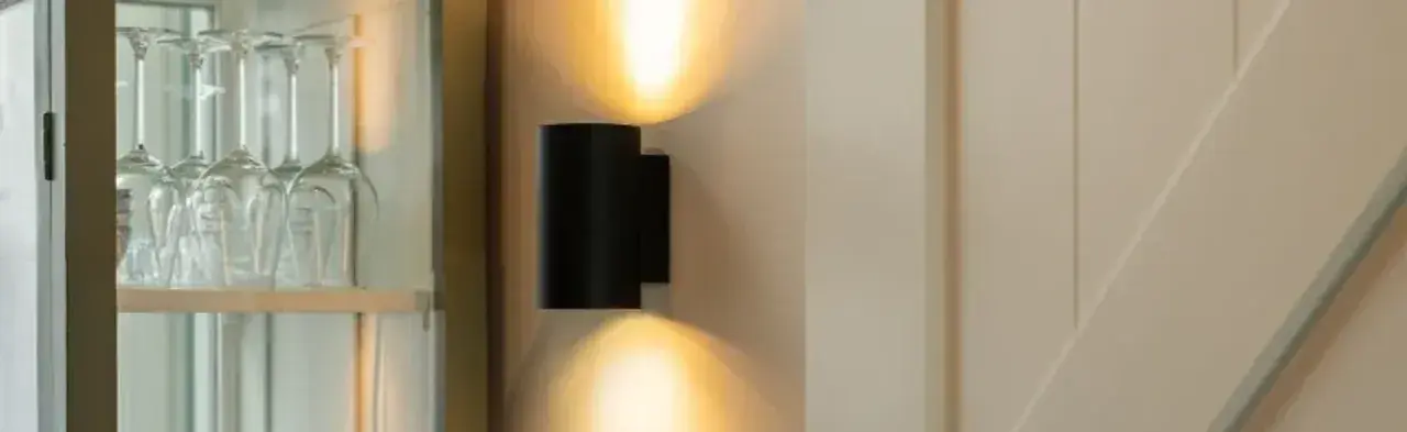 Lámpara de pared original negra, proyectando luz cálida hacia arriba y abajo. Copas de cristal en un armario a la izquierda.