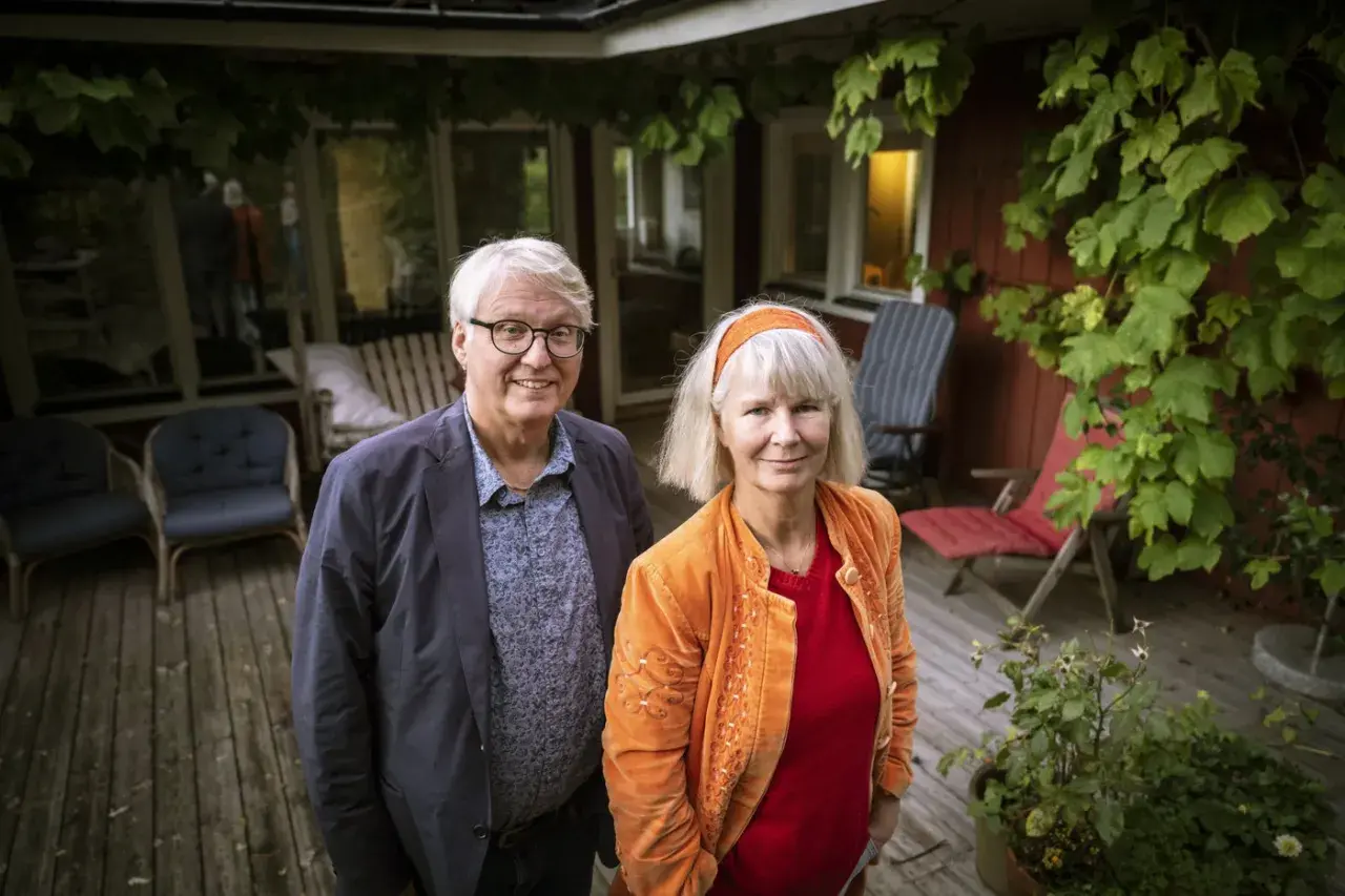 Ett par står på en altan. Mannen har glasögon och en blå kavaj. Kvinnan har ett orange hårband och en orange jacka. De ser ut som en del av amanda romare familj.