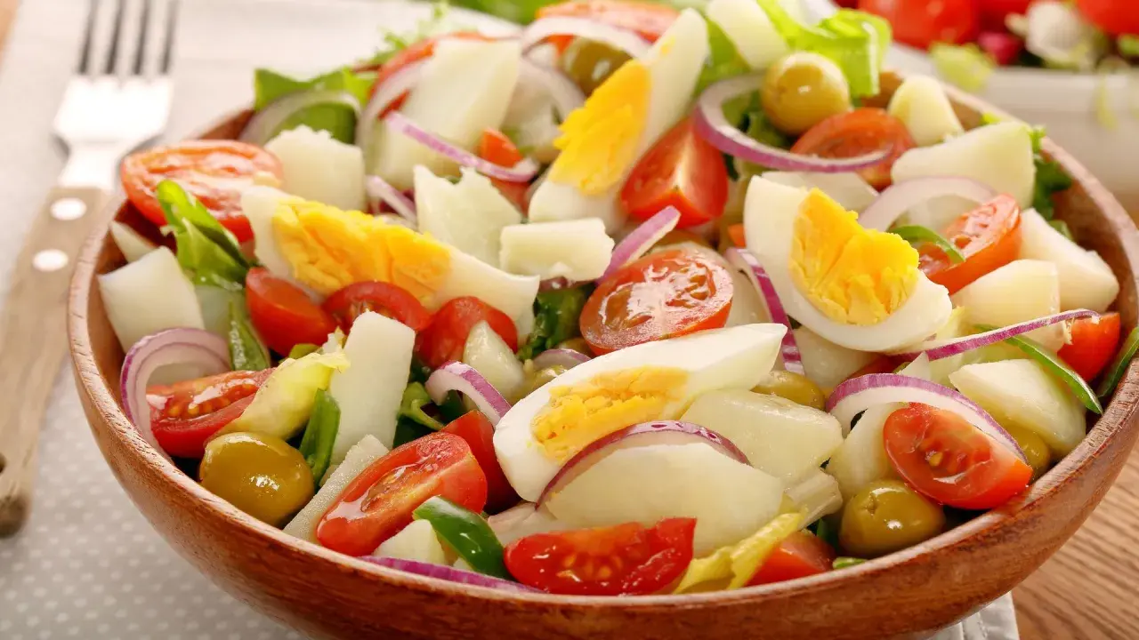 Deliciosa ensalada de patata con huevo duro, tomate, cebolla morada y aceitunas verdes en un cuenco de madera.