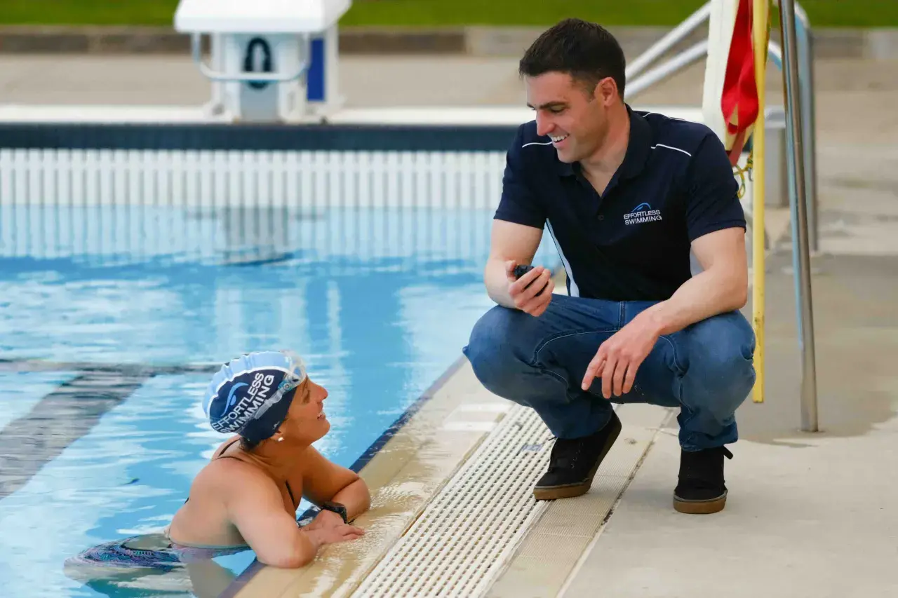 A swim coach crouches by the pool, holding a stopwatch, to discuss swim technique with a swimmer.