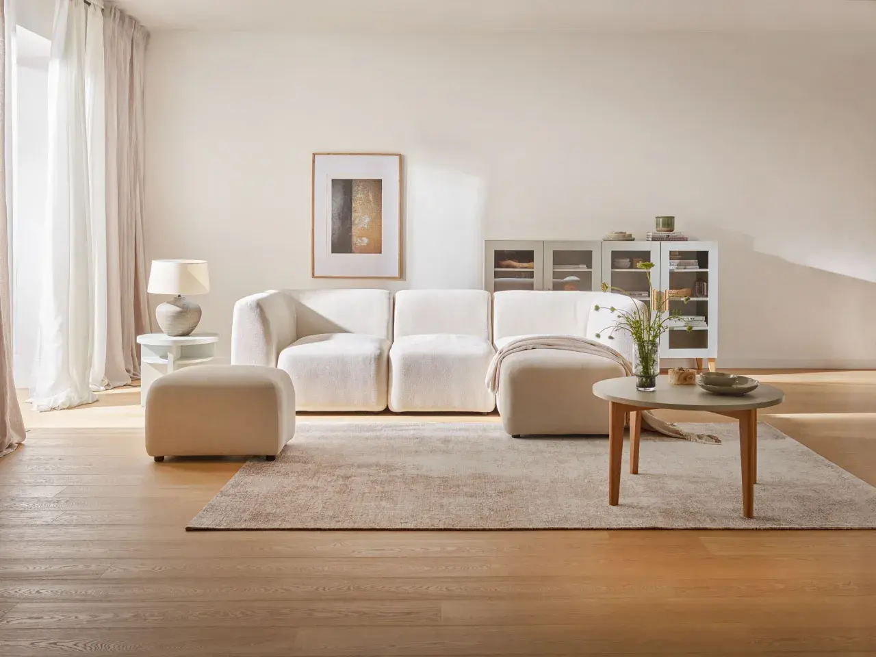 Przytulny salon z kanapą i pufą w odcieniach beżu farby. Jasne zasłony, drewniana podłoga i subtelne dekoracje tworzą spokojną atmosferę.