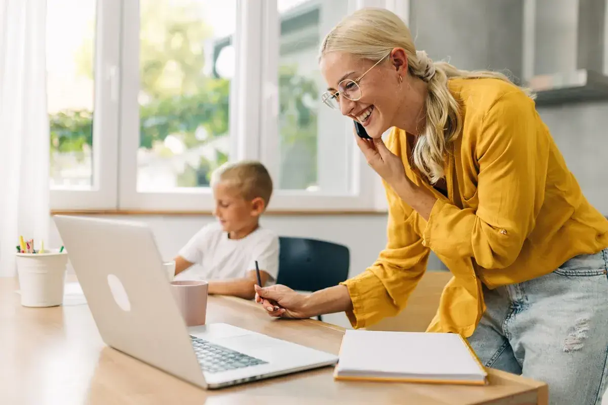 Uśmiechnięta kobieta rozmawia przez telefon, pracując przy laptopie, podczas gdy jej syn rysuje obok. Odpowiedź na pytanie: czy na urlopie wychowawczym można pracować.