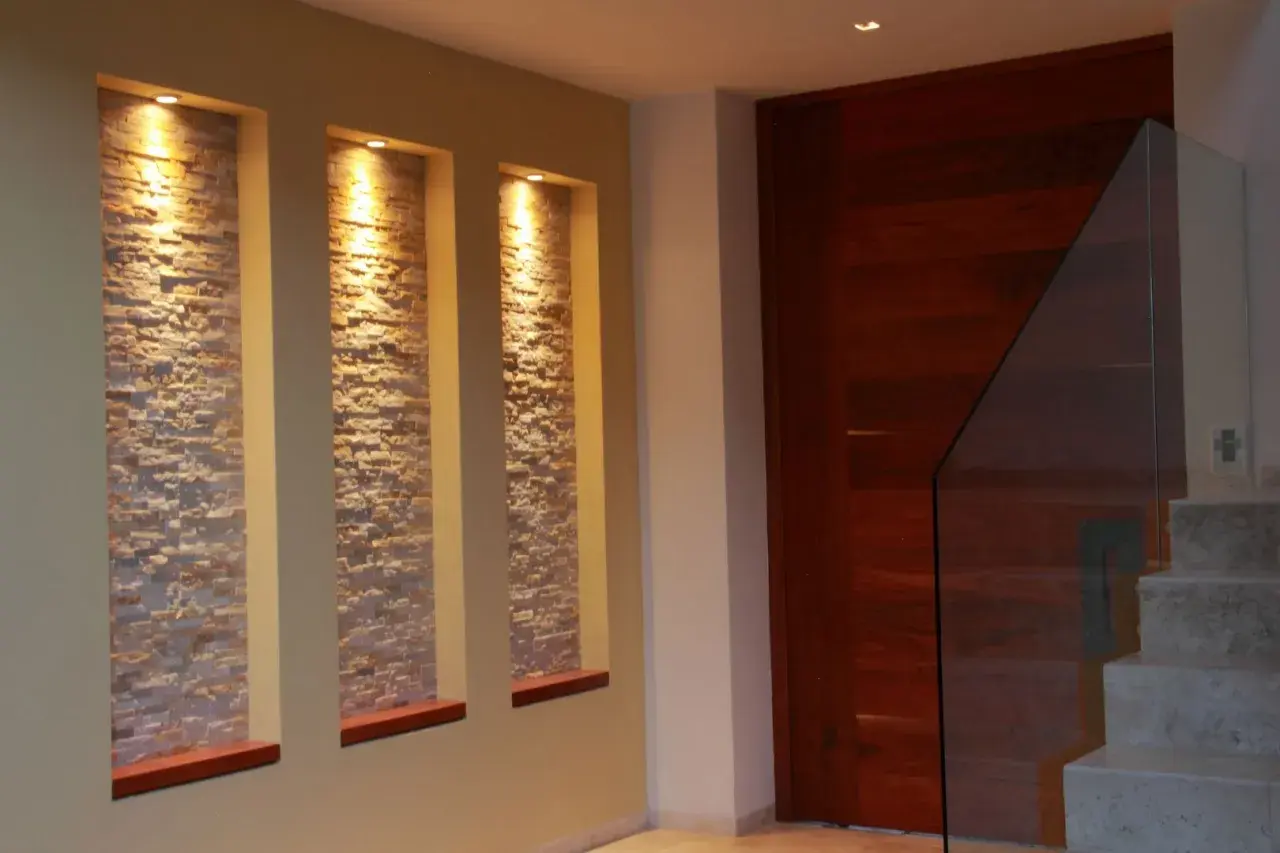 Tre nicchie in cartongesso illuminate da faretti, con rivestimento in pietra, creano un punto focale moderno accanto a una porta in legno e una scala con parapetto in vetro.