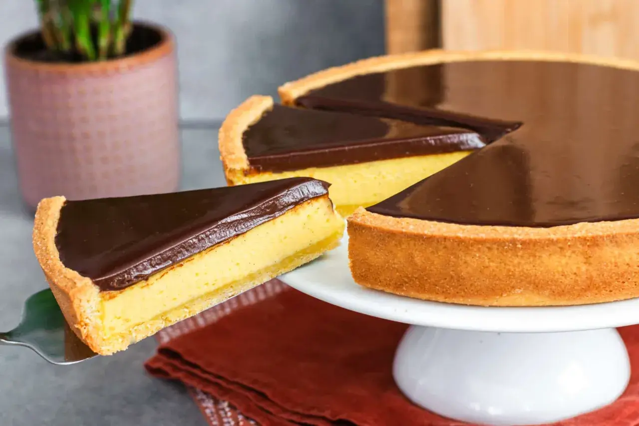 Una fetta di torta Susanna, con crema gialla e copertura al cioccolato fondente, viene sollevata da una tortiera su un'alzata bianca.