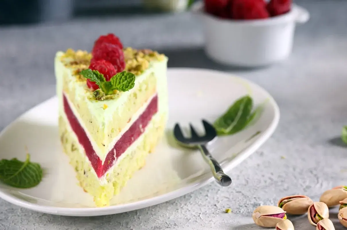 Pyszny tort pistacjowo malinowy z malinami i miętą na talerzu. Obok widoczne są pistacje.