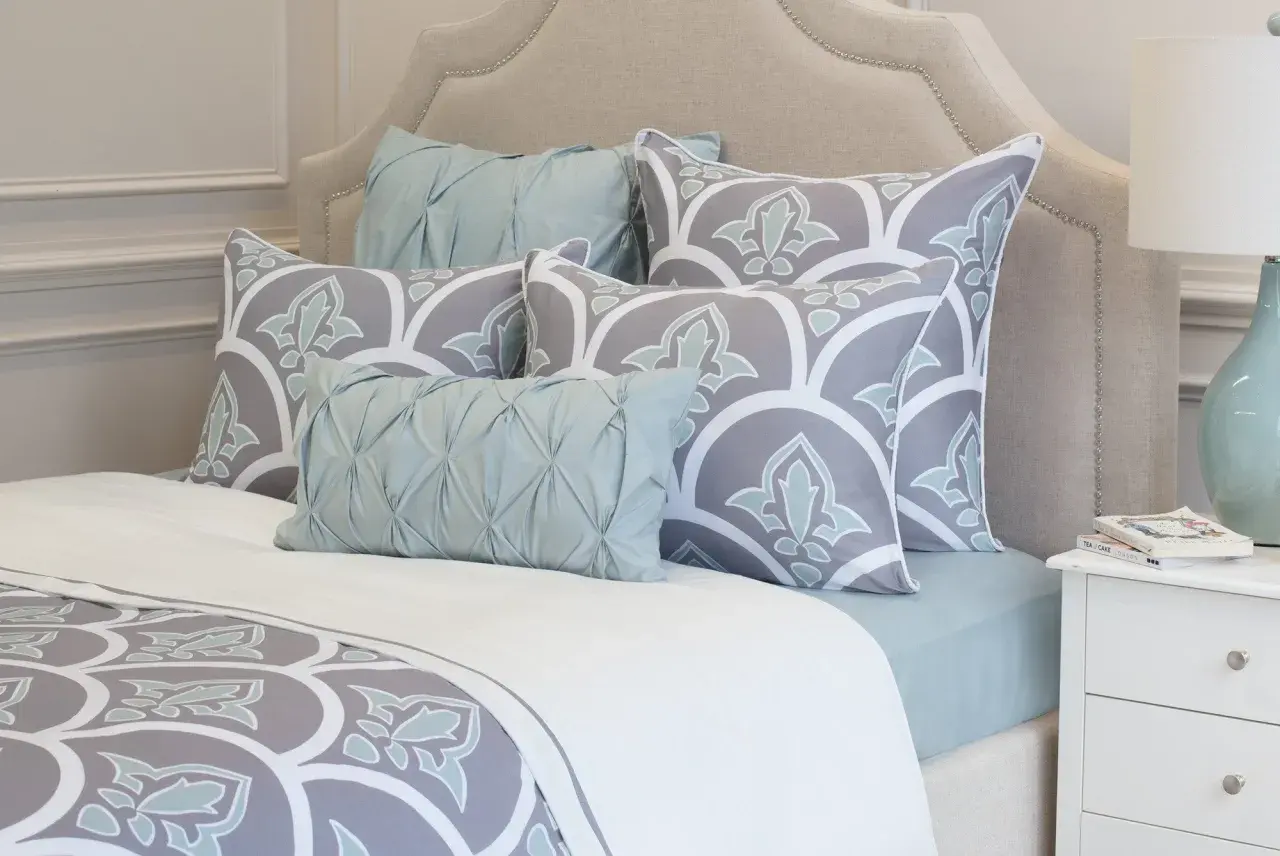 A bed with a tufted headboard, featuring large square pillows with a grey and white floral pattern, and smaller light blue accent pillows.