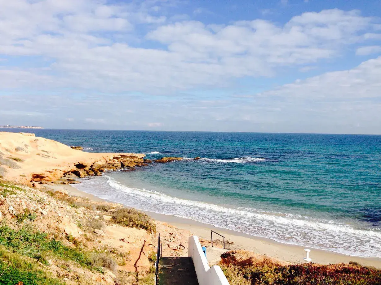 Escaleras descienden a la playa rocamar, con arena dorada, rocas y mar azul bajo un cielo nublado.