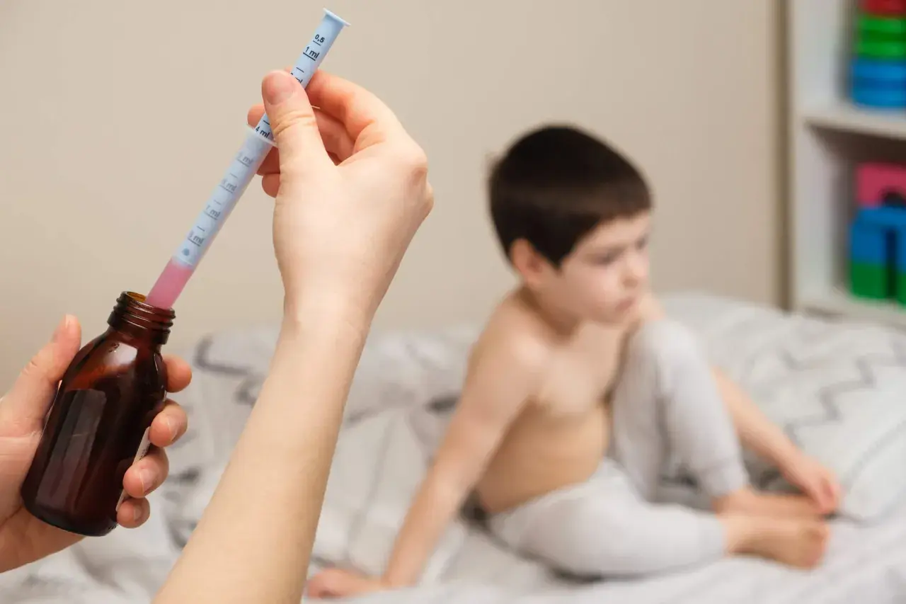 Mano preparando jeringa con ibuprofeno para un bebé. Niño en cama al fondo.