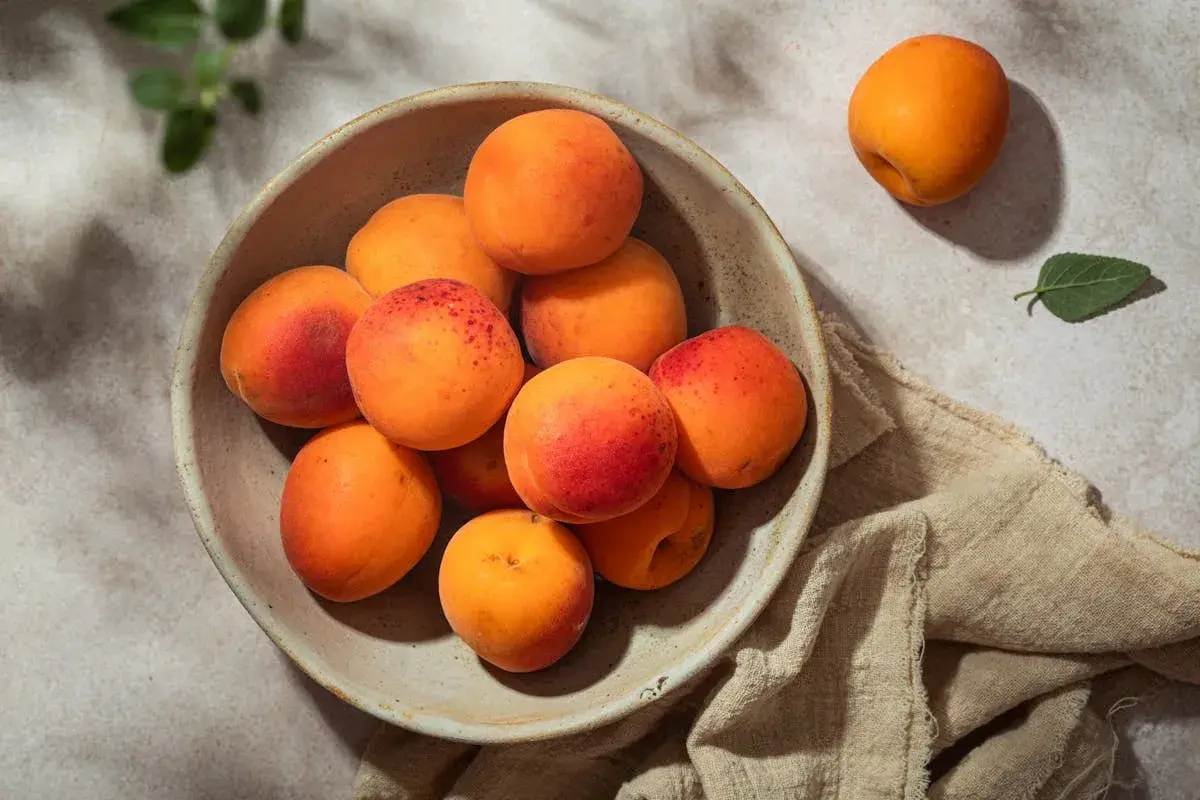 Un bol rempli d'abricots juteux, annonçant la belle **période abricot**. Un fruit isolé repose à côté, sur un tissu beige.