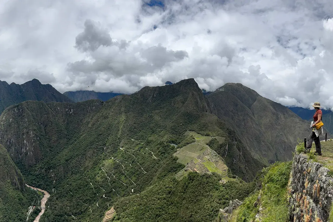 Blick auf Machu Picchu und Huayna Picchu. Eine Frau steht auf einem Felsvorsprung und blickt auf die grüne Berglandschaft.