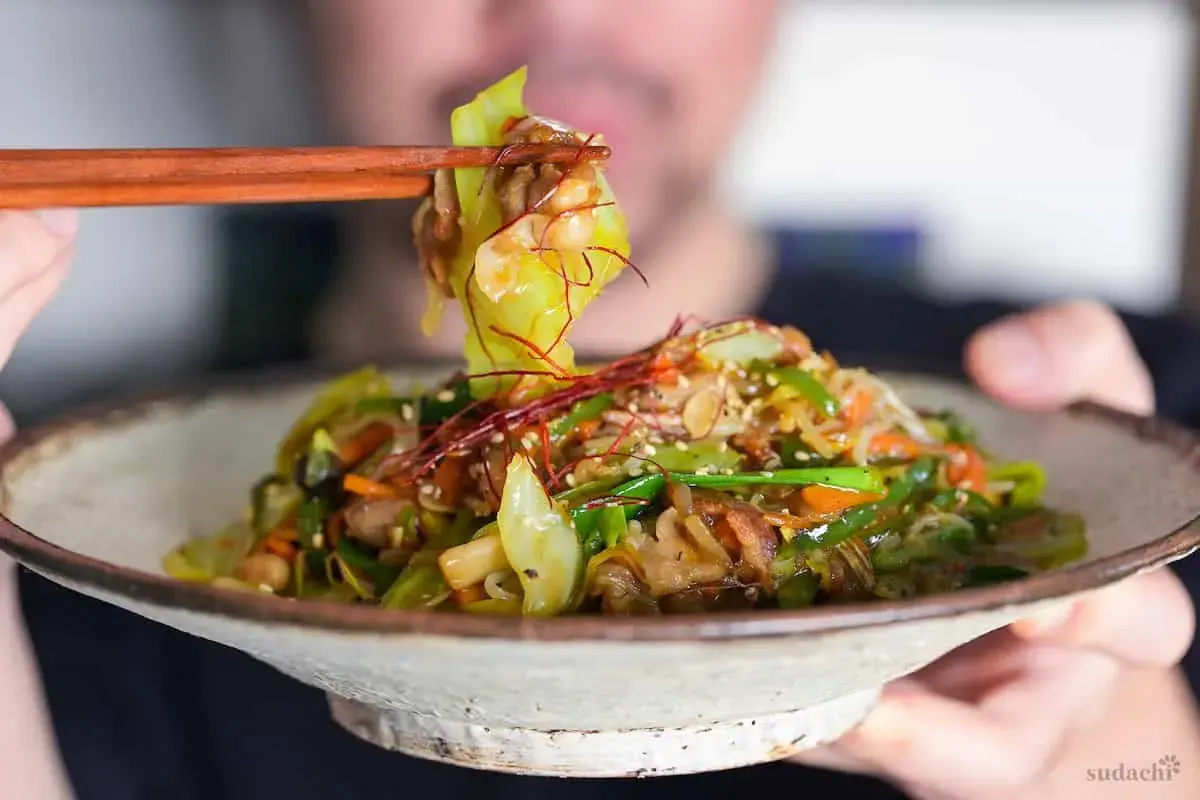 A person uses chopsticks to lift a bite of a colorful Japanese stir fry, featuring cabbage, carrots, and peppers, from a rustic bowl.