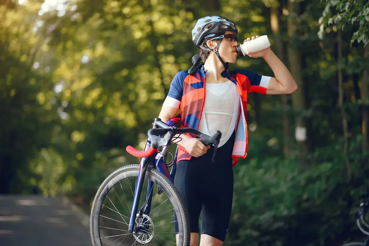 Kolarz w stroju sportowym pije wodę z bidonu, dbając o odpowiednią dietę kolarza. W tle zielony las.