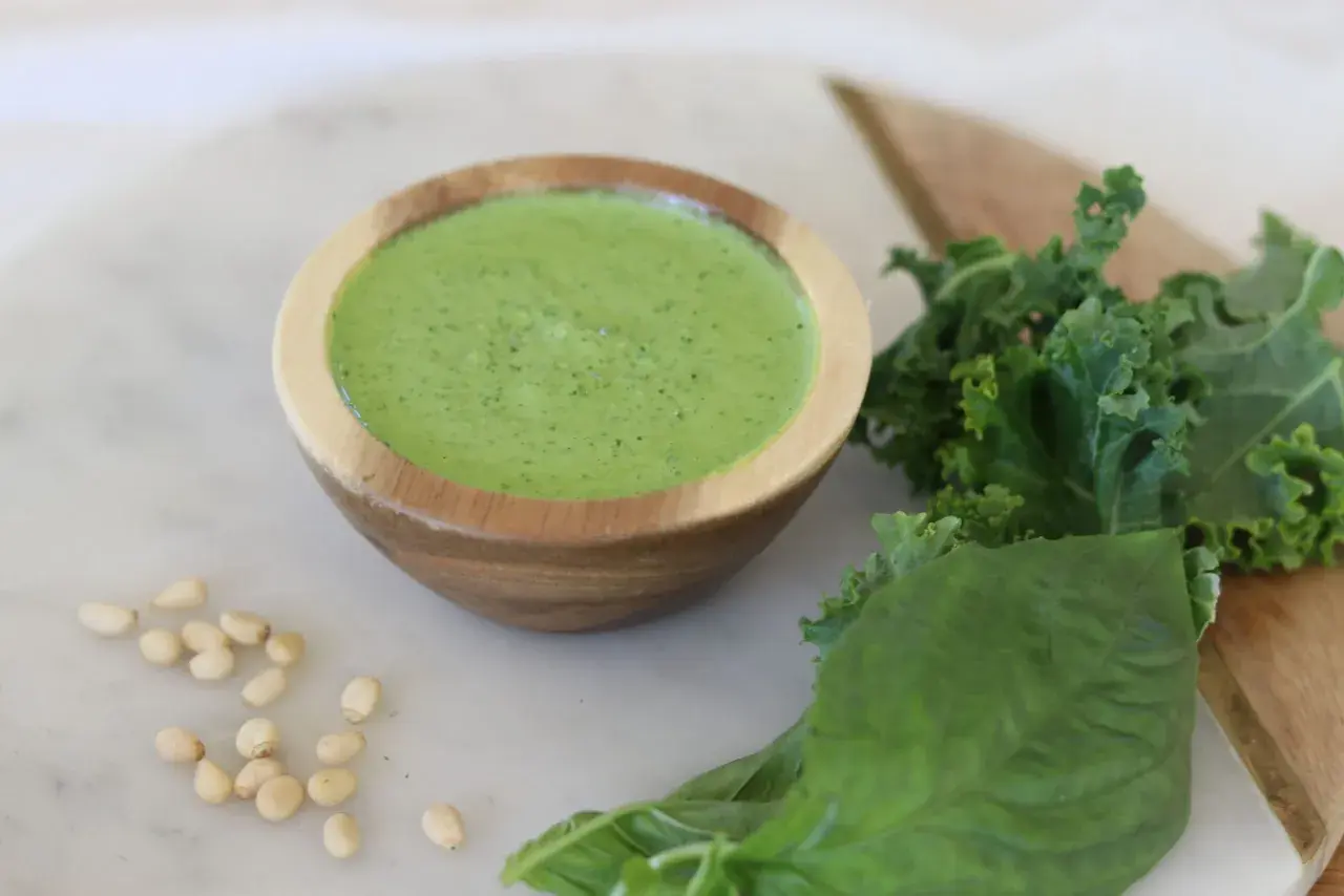 Un cuenco de madera contiene un vibrante pesto de kale, acompañado de hojas frescas de kale, albahaca y piñones.