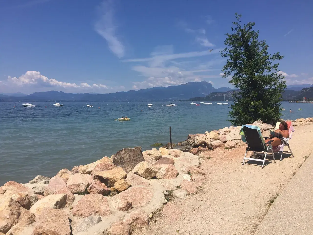Una donna legge su una sedia a sdraio sulla riva rocciosa di Cisano, Gardasee. Barche galleggiano sull'acqua calma con montagne sullo sfondo.