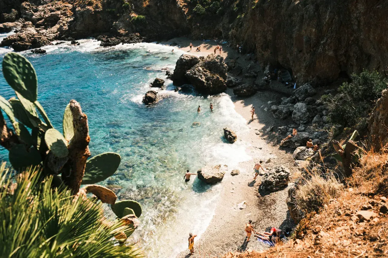 A secluded beach in Zingaro, Sicily, with crystal-clear turquoise water, rocky cliffs, and sunbathers enjoying the Mediterranean.