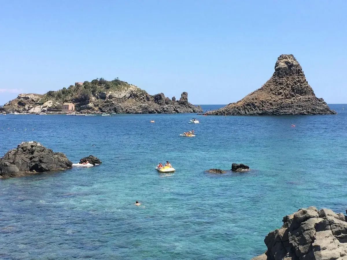 Vue sur la mer bleue et les rochers volcaniques d'Aci Trezza, Sicile. Des bateaux et des baigneurs profitent du soleil.
