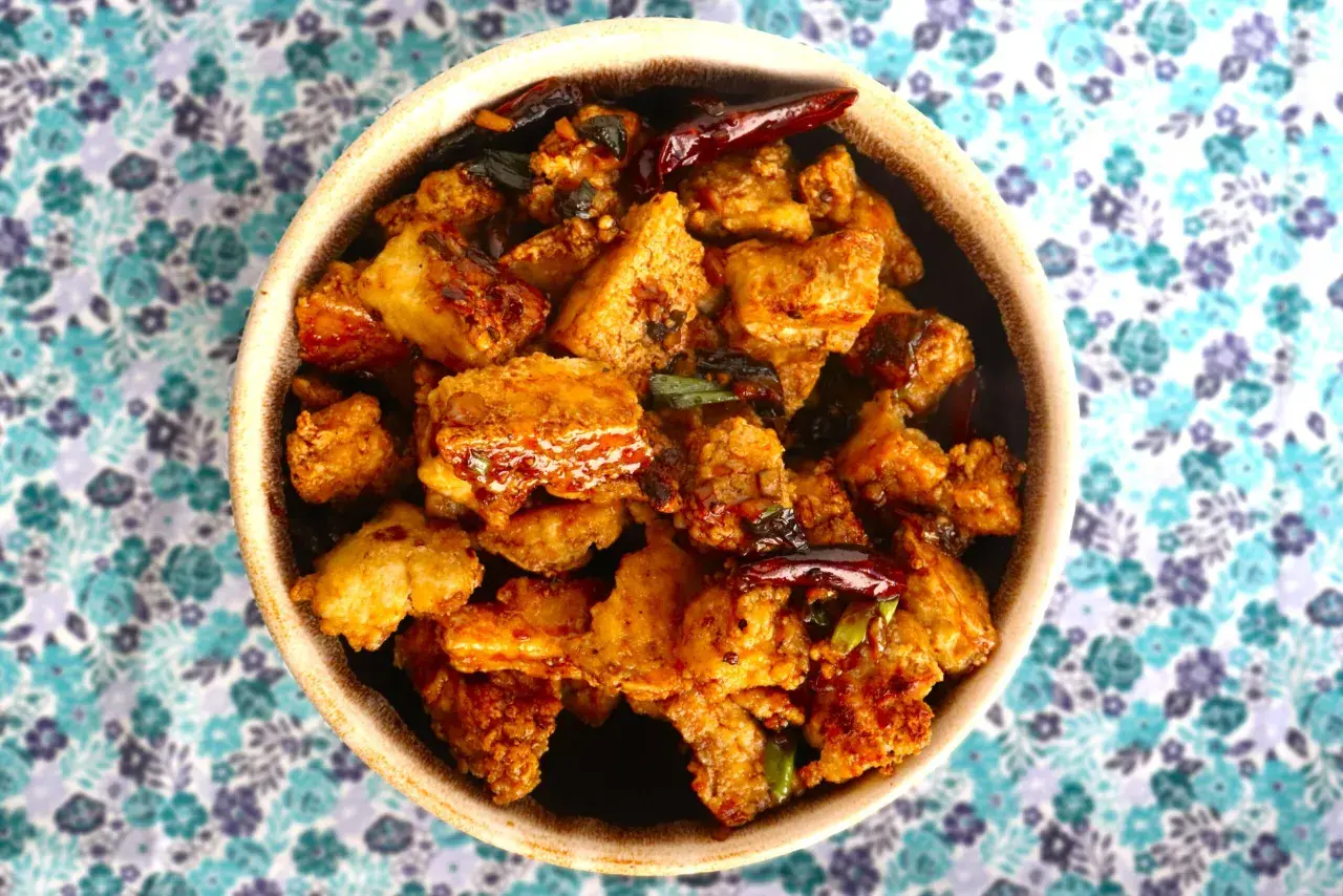 Crispy, golden-brown pieces of ice tofu are coated in a savory sauce with chili peppers and green onions, served in a bowl on a floral background.