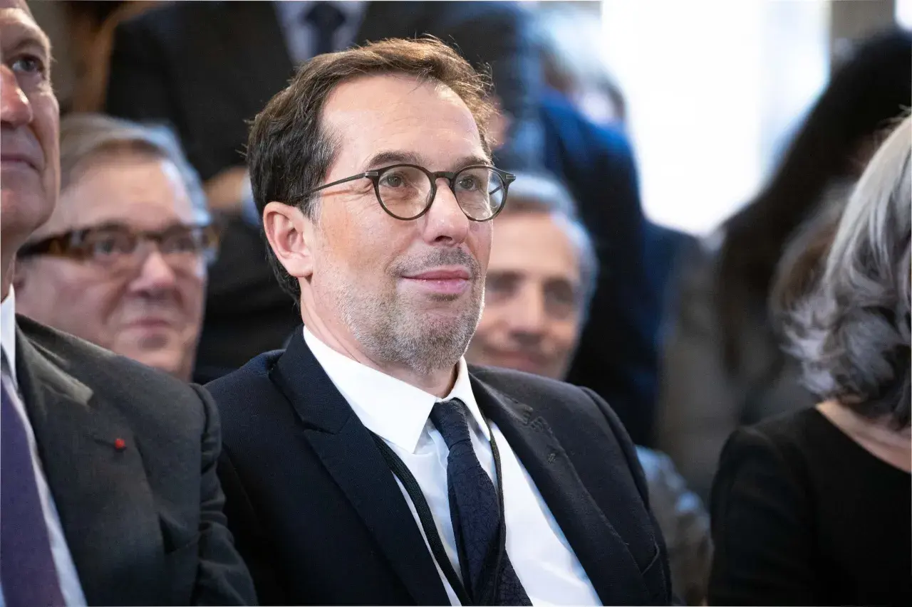 Un homme avec des lunettes, en costume noir, souriant.
