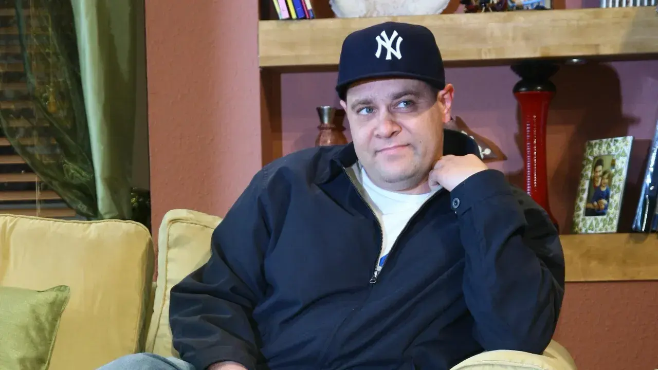 Mężczyzna w czapce z daszkiem Yankees, w wieku, który można by określić jako dojrzały, siedzi na kanapie.