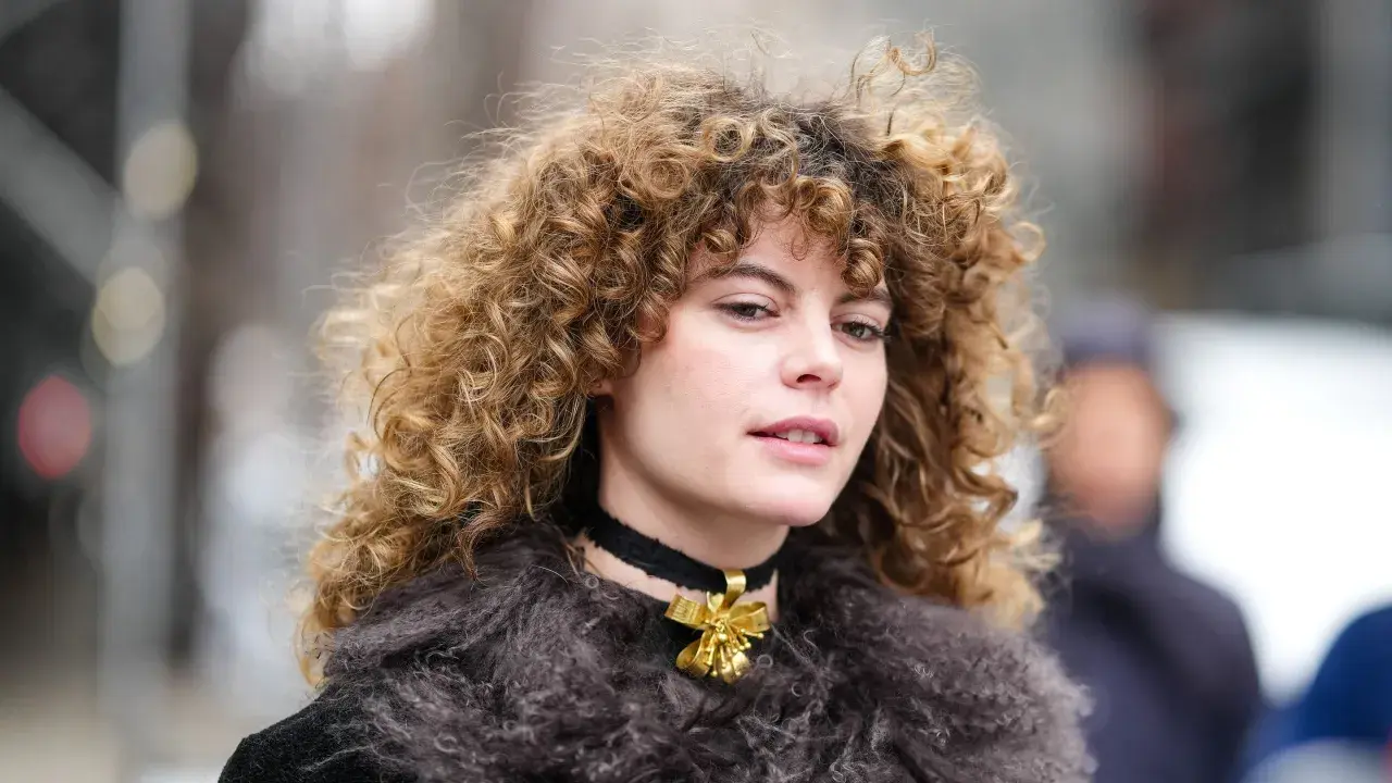 Eine Frau mit voluminösen, krausen Haaren trägt einen Pelzmantel und eine Halskette mit einer goldenen Schleife.