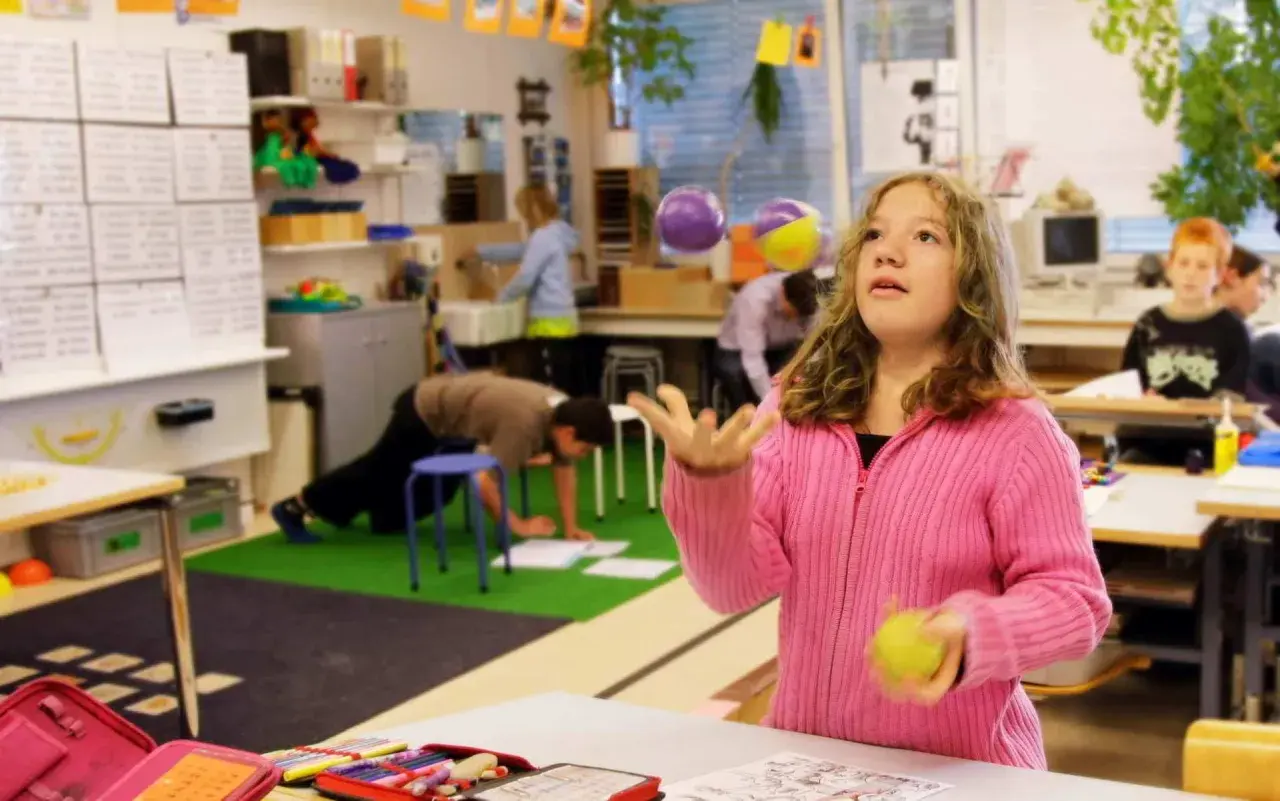 Mädchen jongliert mit Bällen im Klassenzimmer, während andere Kinder lernen. Ein Beispiel für bewegtes Lernen.