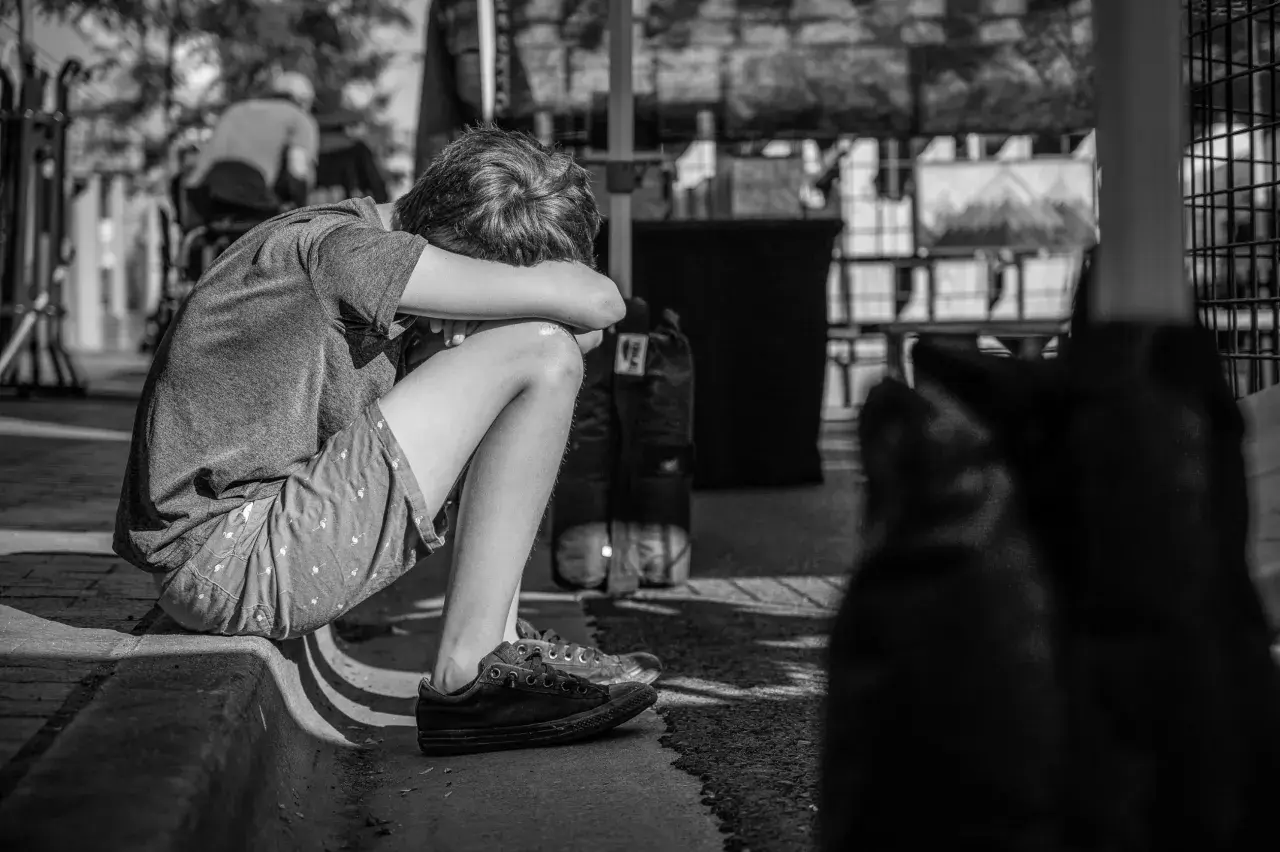 Chłopiec skulony na chodniku, zamyślony. Leczenie psychiatryczne a opieka nad dzieckiem to trudne wyzwania dla rodziny.