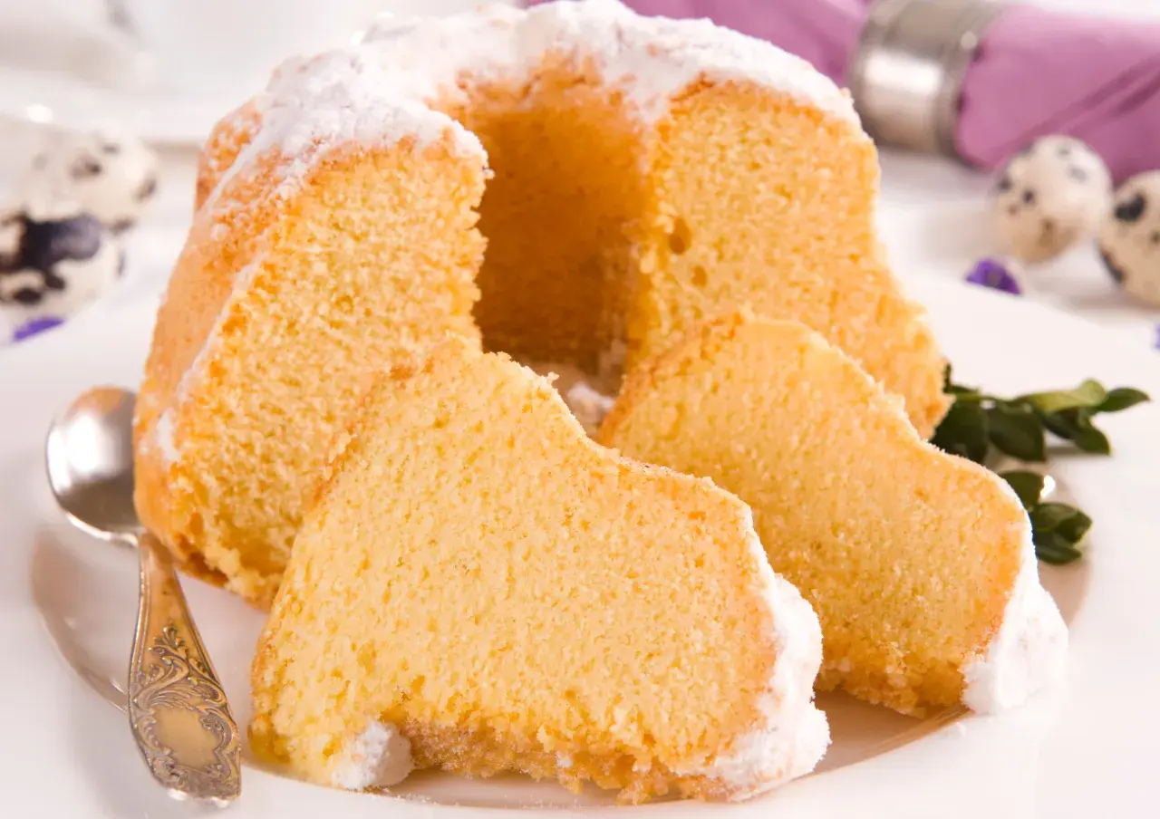 Puszysta babka z mascarpone, oprószona cukrem pudrem, z kilkoma kawałkami na talerzu. Obok leży łyżeczka.