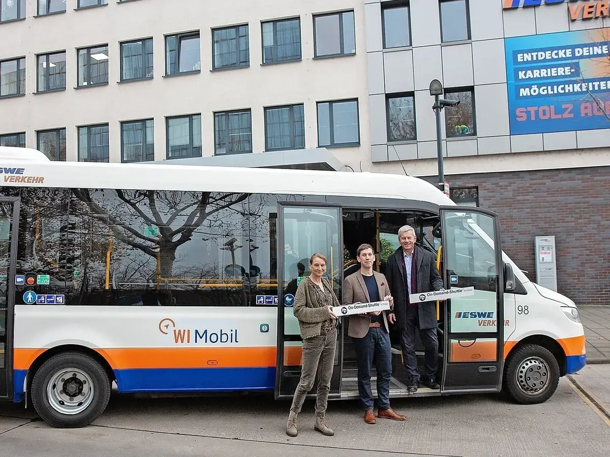 Drei Personen stehen vor einem weißen Kleinbus mit der Aufschrift "WI Mobil". Sie halten Schilder mit der Aufschrift "On-Demand-Shuttle".