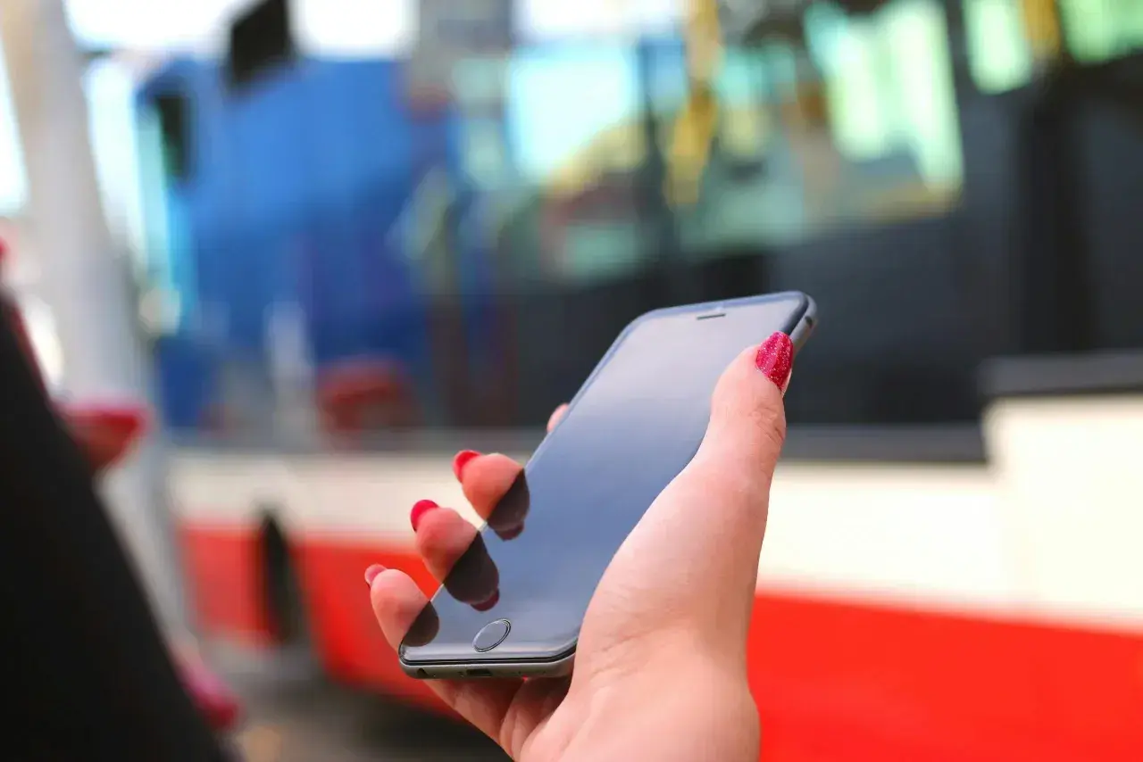 Dłoń z czerwonymi paznokciami trzyma smartfon, na którym można kupić bilety ZTM Warszawa. W tle widać autobus.