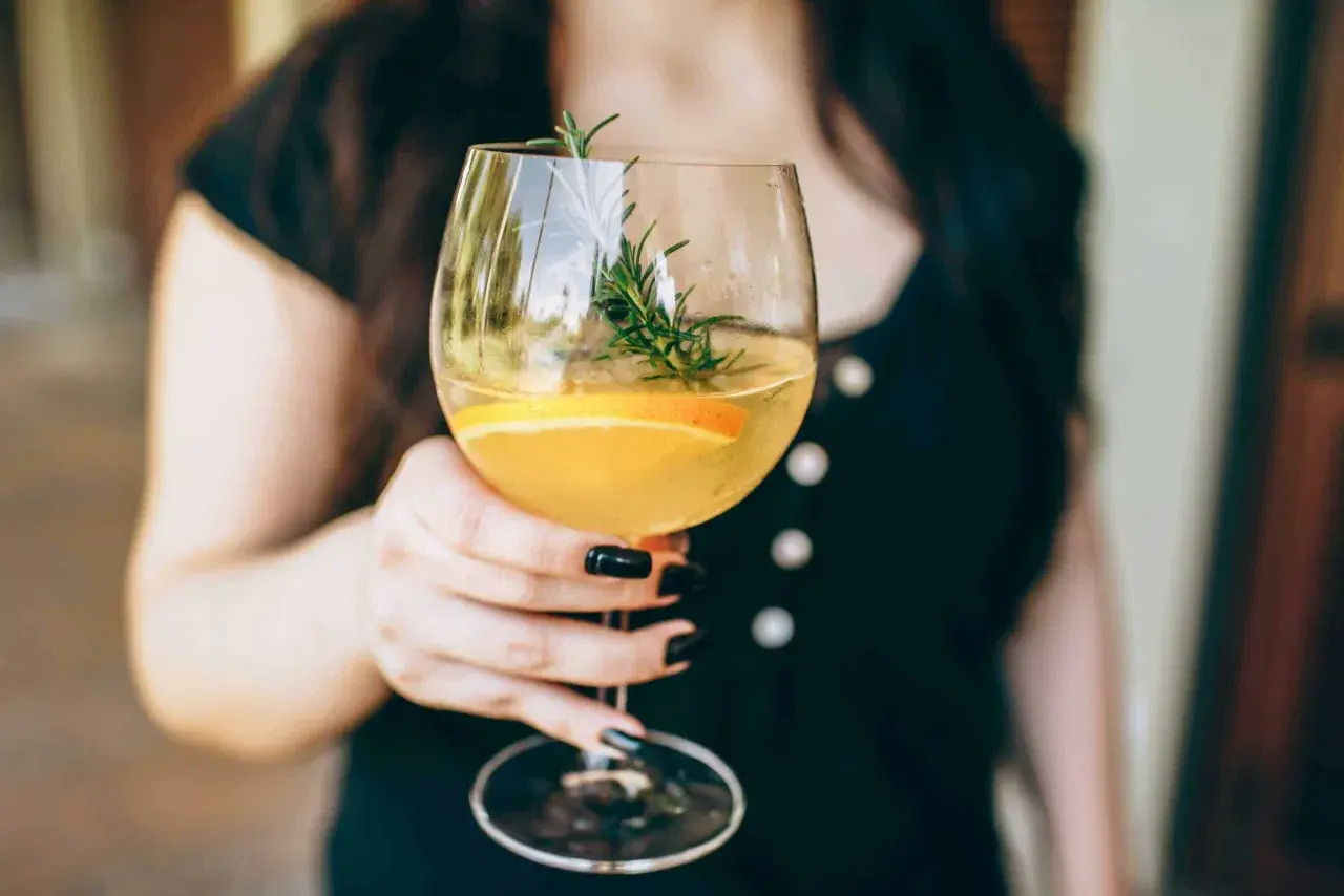 En person håller ett glas med en uppfriskande drink med vitt vin, garnerad med en apelsinskiva och en rosmarinkvist.