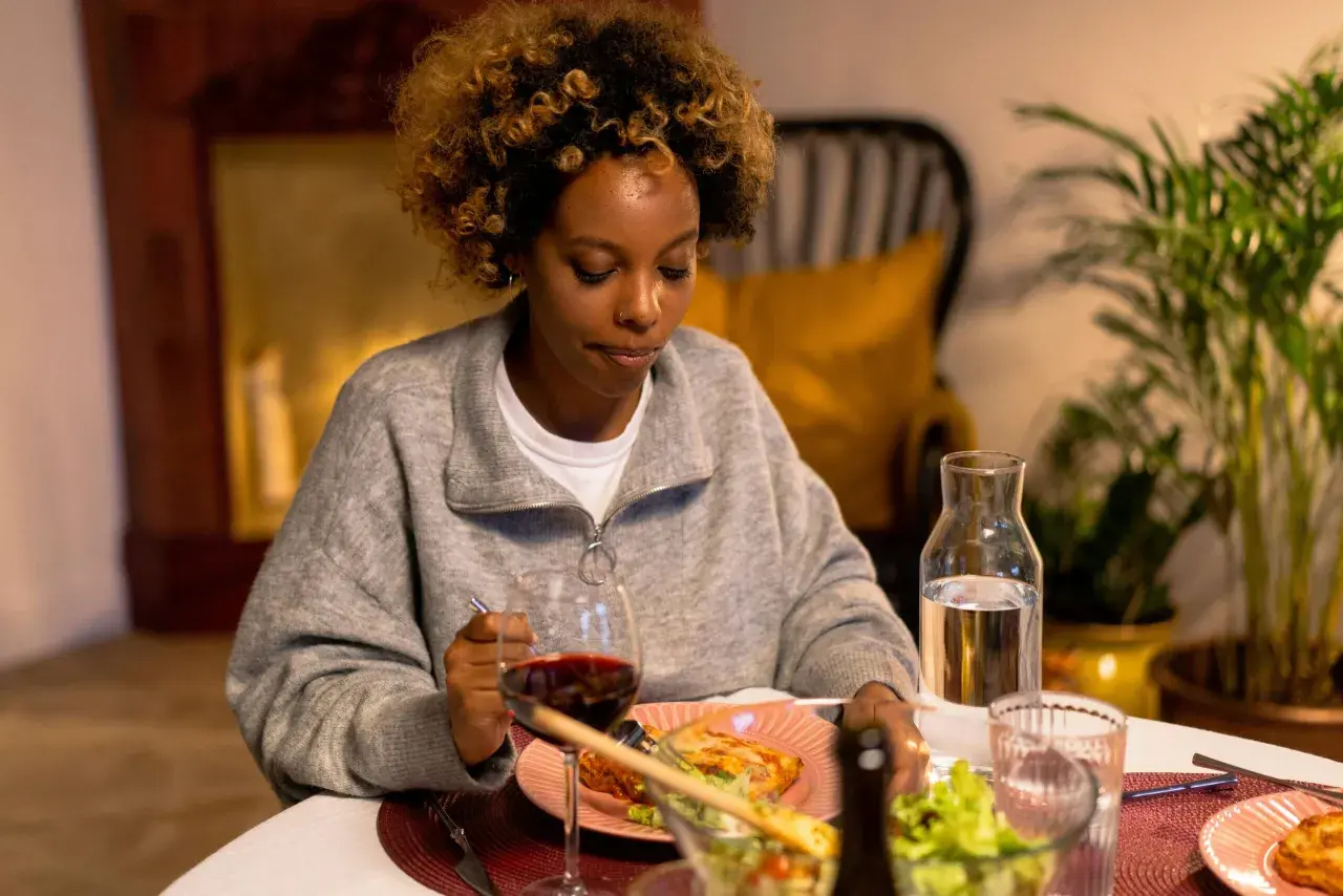 Mujer con cabello rizado mira su plato de comida, pero no come porque no tengo hambre. Hay vino y ensalada en la mesa.