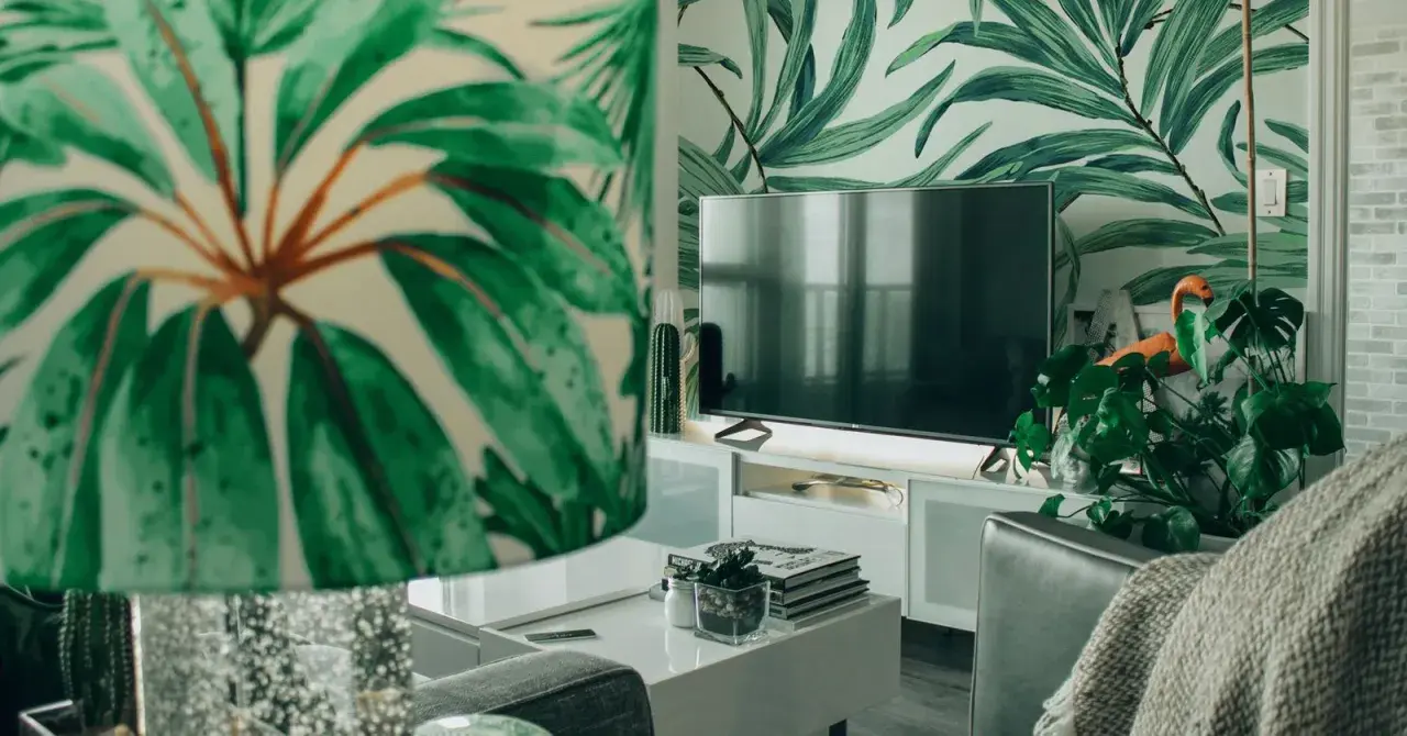 Sala de estar con papel tapiz de hojas tropicales. Muebles DM blancos y un televisor moderno.