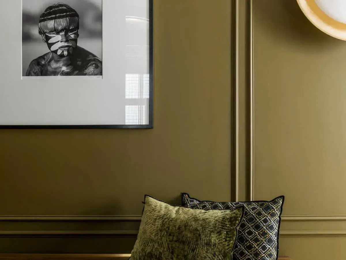 Un pan de mur vert olive, orné d'un portrait encadré d'un jeune homme au visage peint, et de coussins décoratifs.