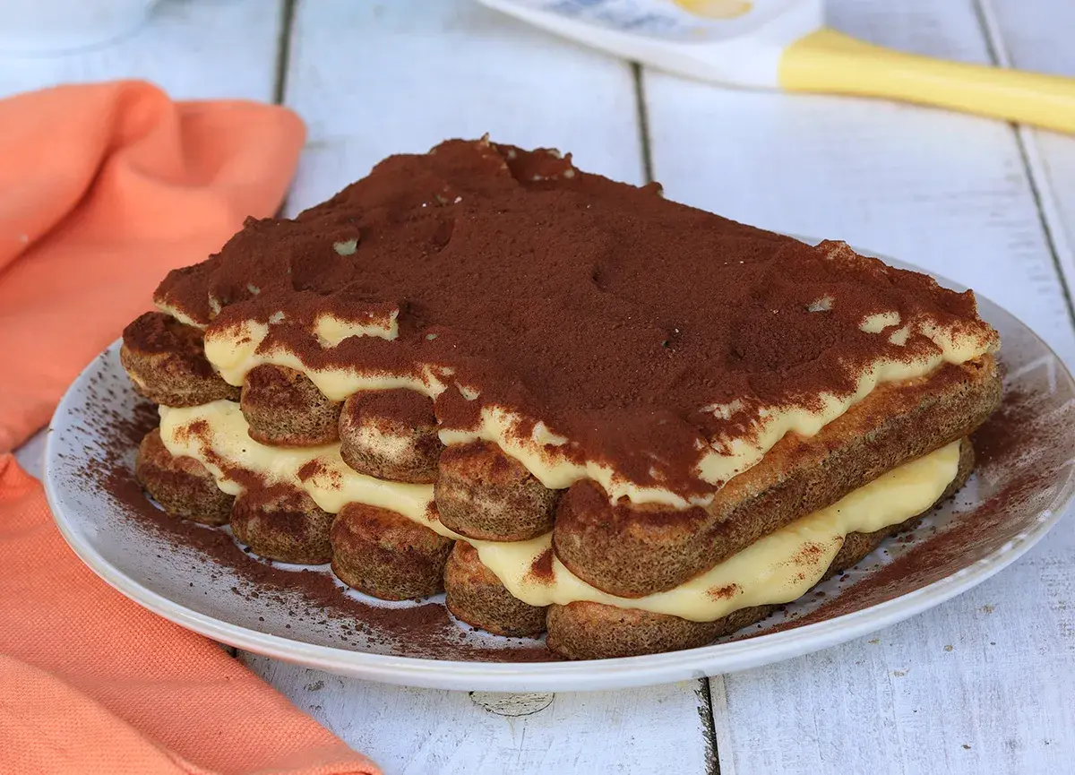 Tiramisù con crema pasticcera, strati di savoiardi e cacao in polvere su un piatto bianco, con un tovagliolo arancione sullo sfondo.