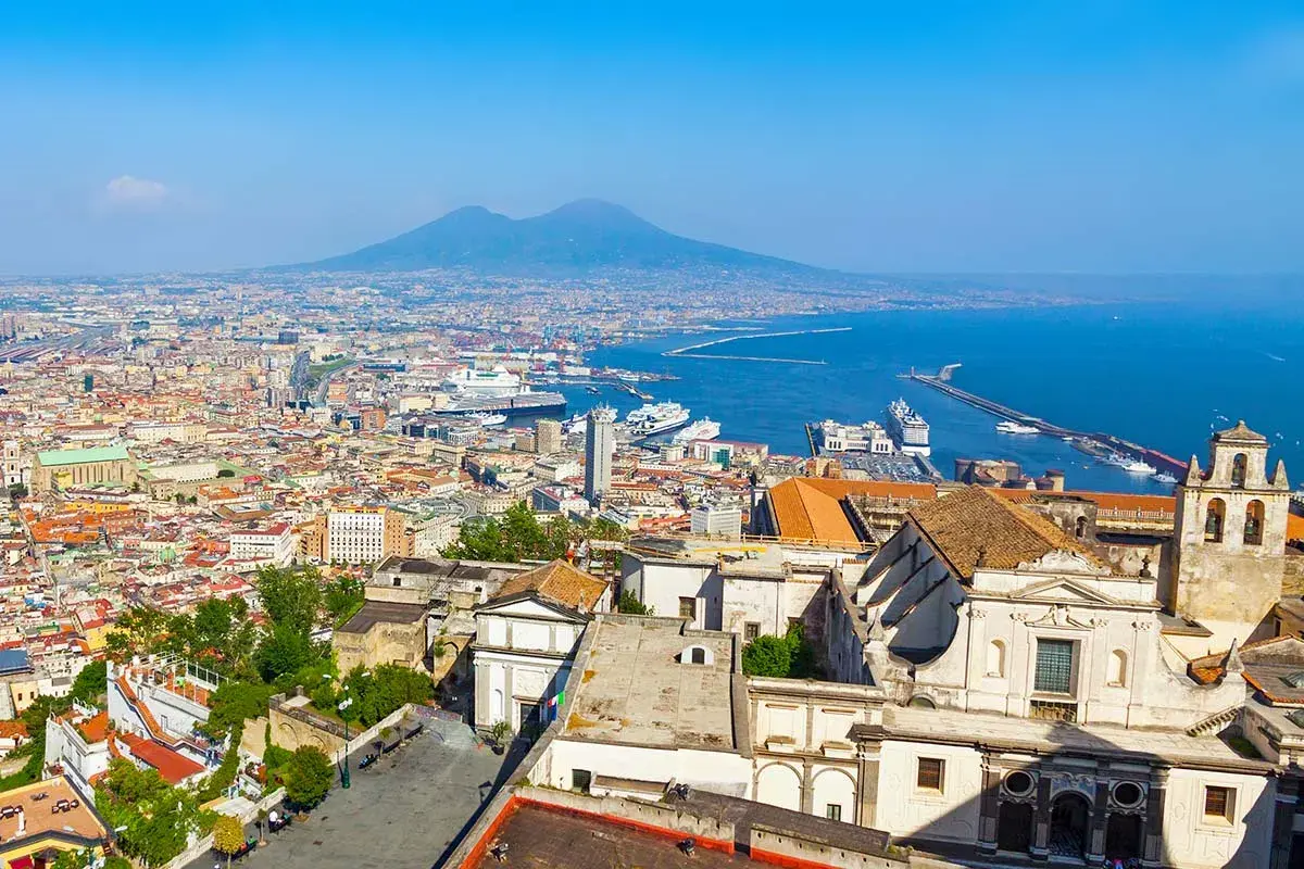 A panoramic view of Naples, Italy, with Mount Vesuvius looming in the distance. The city's historic buildings and bustling port suggest that Naples is indeed nice.